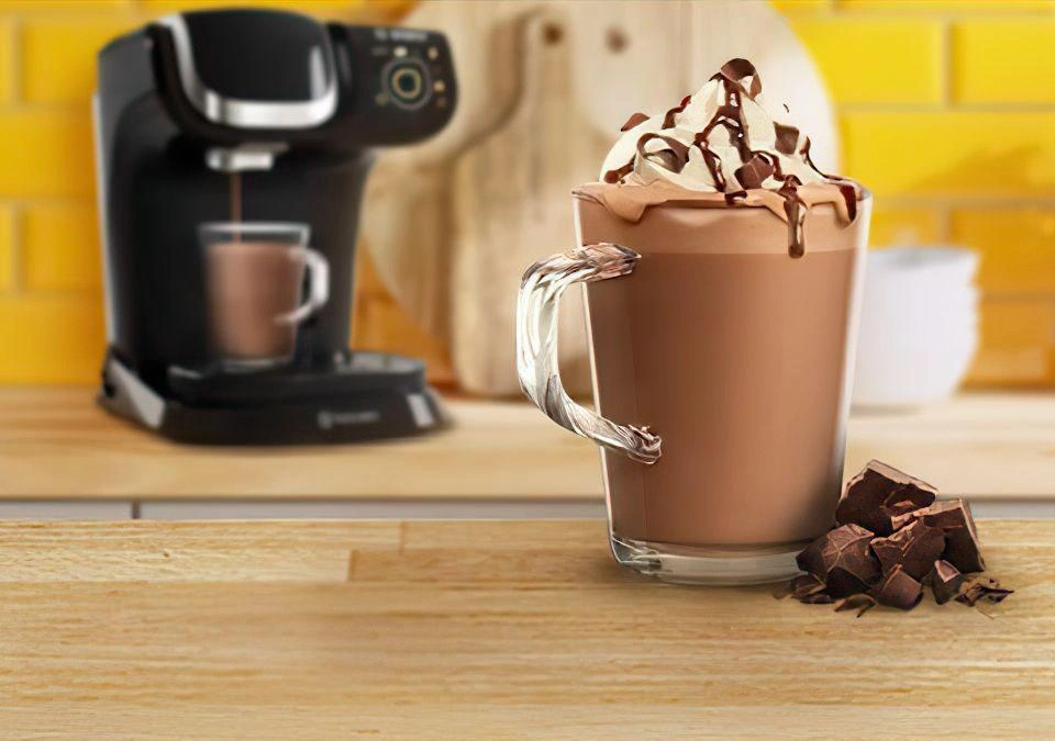 A steaming glass mug of hot chocolate sits enticingly on the table, topped with a swirl of whipped cream and delicate chocolate shavings. Nestled beside it is a TASSIMO coffee pod, promising an easy indulgence in rich flavors.