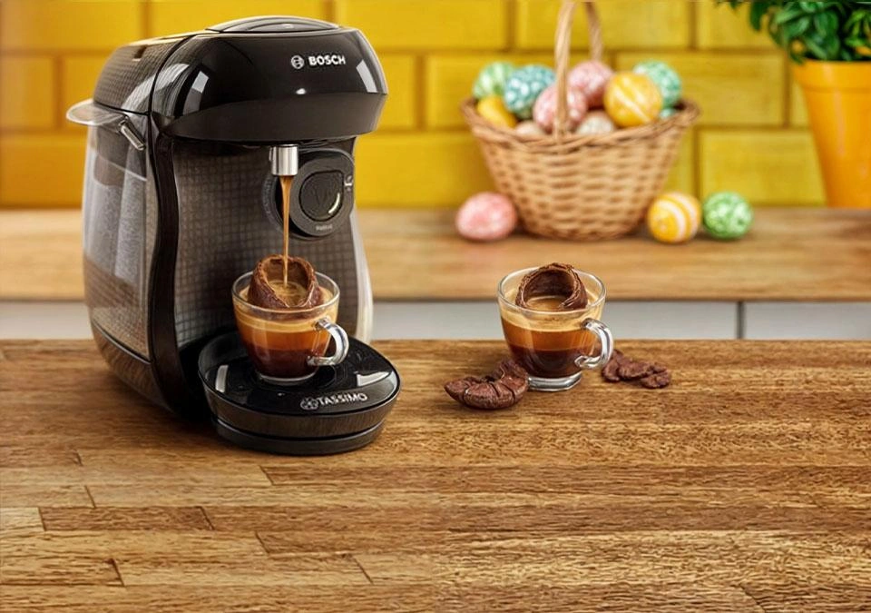A TASSIMO coffee machine brews alongside two espresso cups on a wooden counter, with a basket in the background.