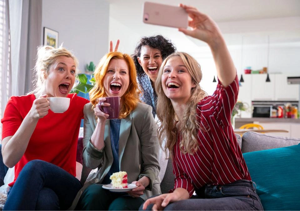 Four women smiling, taking a selfie together in a cozy living room with a TASSIMO coffee machine in the background.