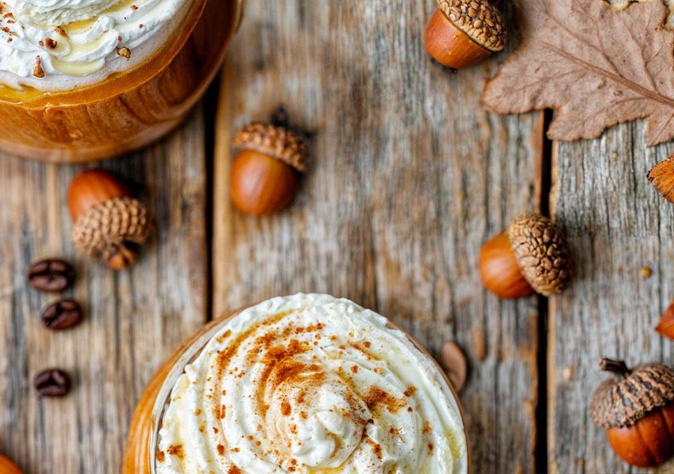 Two TASSIMO pumpkin spice lattes topped with whipped cream sit elegantly on a rustic wood table, surrounded by scattered acorns.