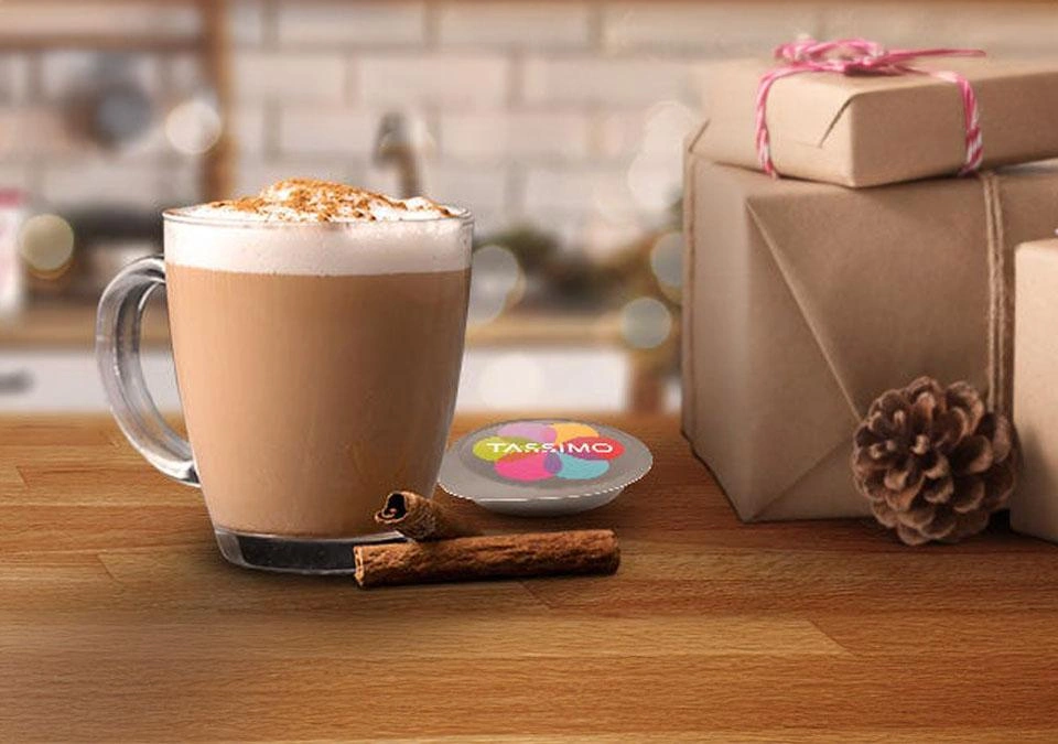 TASSIMO A latte with cinnamon, pinecone, and wrapped gifts on a wooden table.