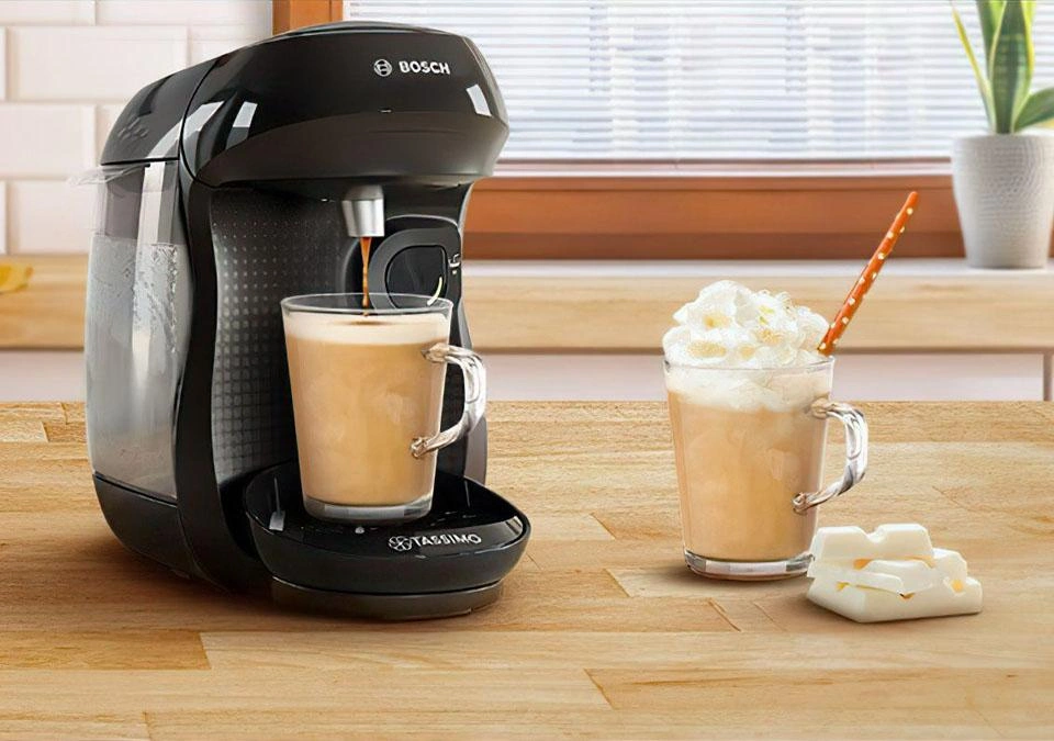 A TASSIMO coffee machine sits on the counter, accompanied by two mugs filled with frothy coffee and topped with white chocolate shavings.