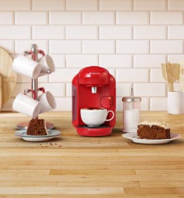 A red TASSIMO coffee maker sits on the counter, surrounded by plates of cake slices and mugs hanging nearby, ready for a cozy gathering.