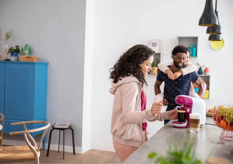A woman brews a cup of TASSIMO coffee in a bright kitchen, smiling as her partner hugs her warmly from behind.