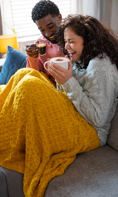 A couple laughing and enjoying TASSIMO coffee on a cozy couch wrapped in a yellow blanket.