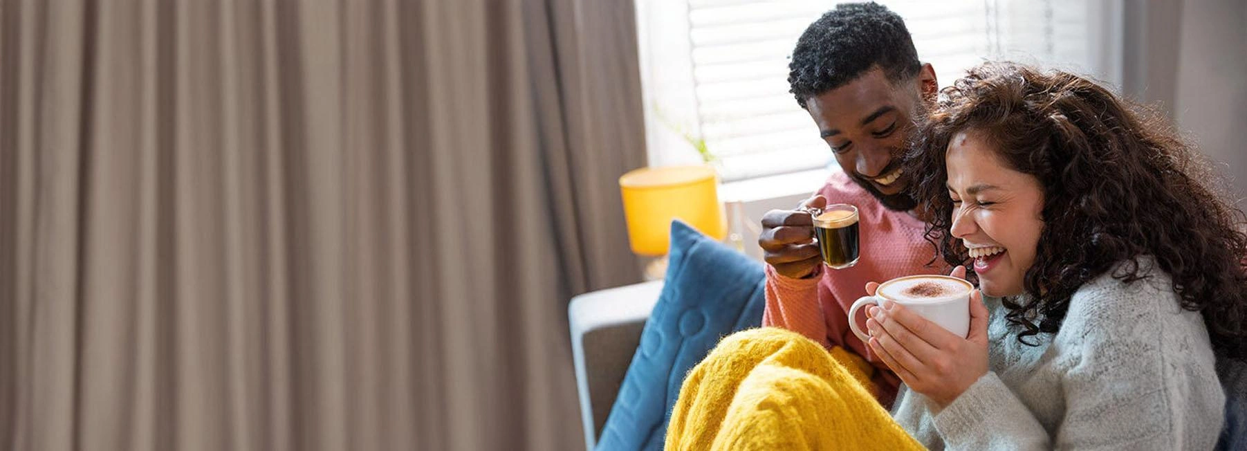 A couple laughing and enjoying TASSIMO coffee under a cozy yellow blanket indoors.