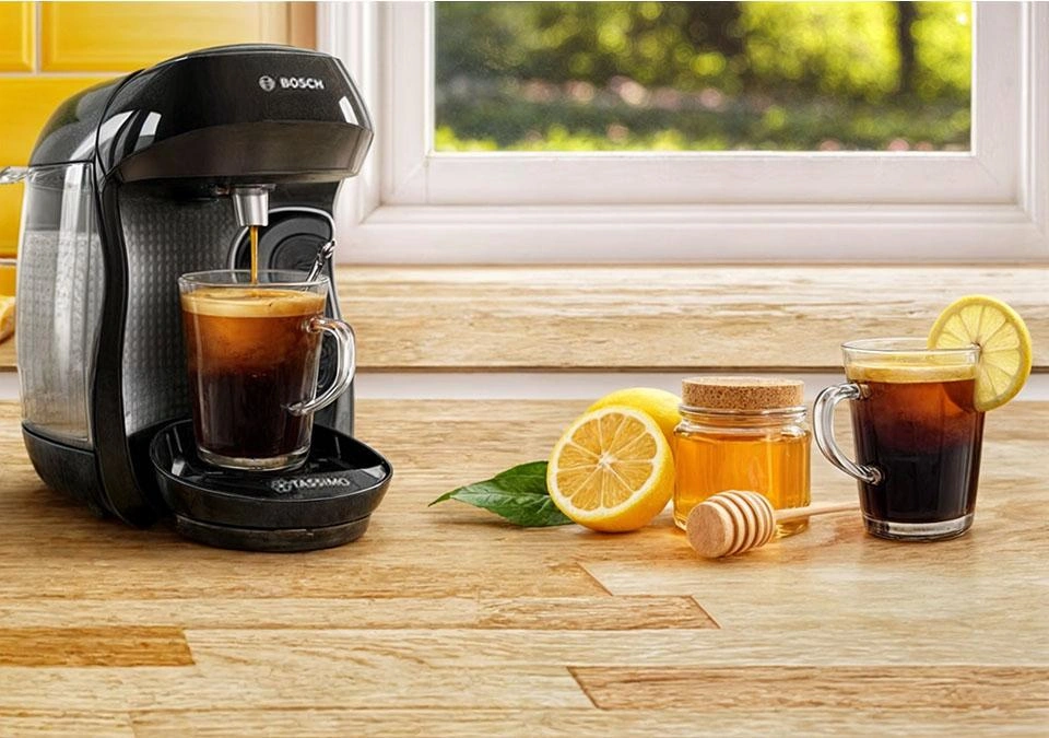 A TASSIMO coffee machine sits on the kitchen counter, with a refreshing iced coffee next to it. Nearby, a jar of honey glistens in the sunlight streaming through the window, offering a serene view outside.