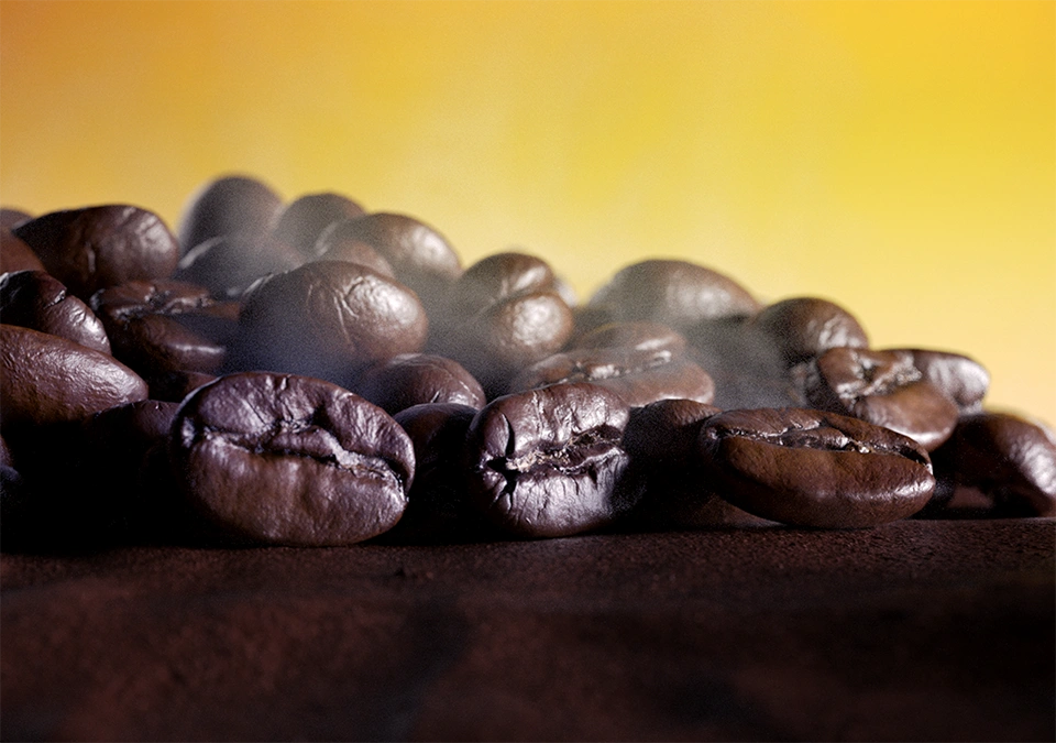 Grains de café L’OR fraîchement torréfiés avec vapeur, sur fond jaune doré.