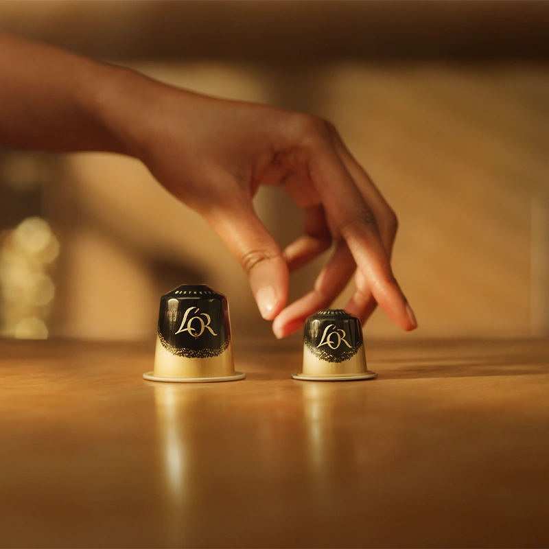 Deux capsules L'OR sur une table dorée : une Ristretto classique et une XXL Ristretto. Une main saisit délicatement la capsule classique.