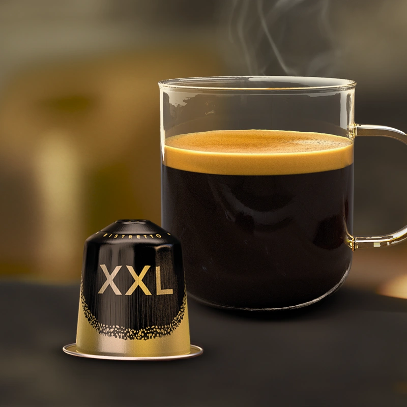 Una taza grande de vidrio llena de café y una cápsula XXL sobre una mesa oscura.