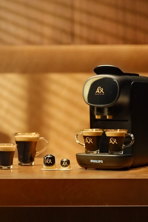 L'OR Sublime black machine with different coffee drinks and capsules on a table.