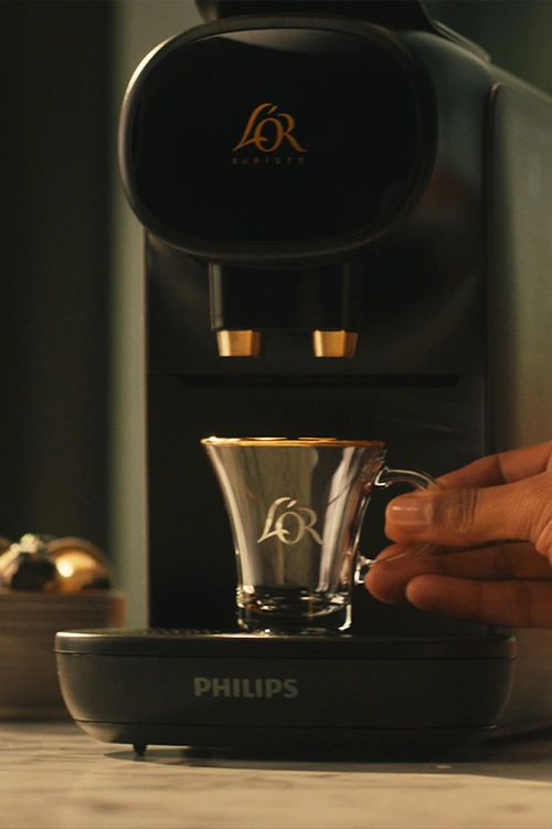 Hand placing a glass L’OR cup on the drip tray below the coffee spouts.