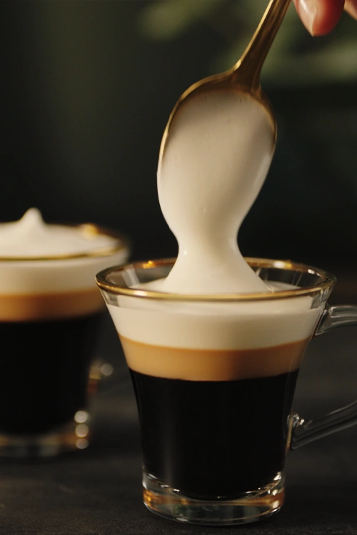 Close-up of a spoon adding thick milk foam onto a layered espresso in a glass cup.