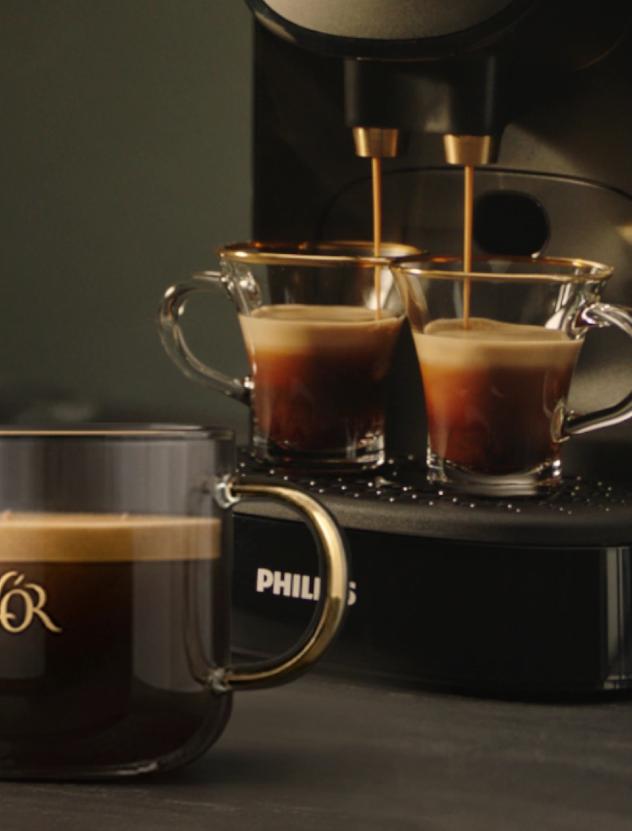 Close-up of a brewed L’OR coffee in a glass mug, with two espressos pouring in the background.