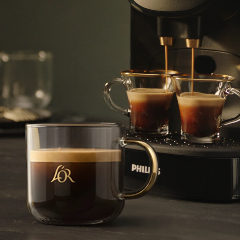 Close-up of a brewed L’OR coffee in a glass mug, with two espressos pouring in the background.