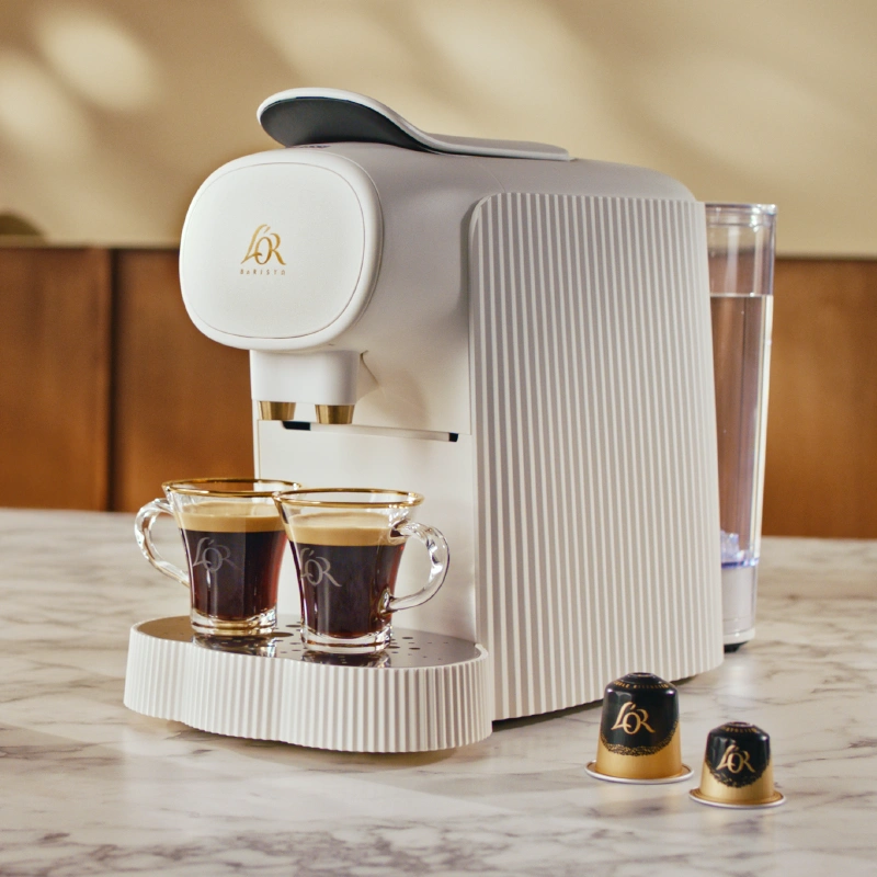 White ribbed L’OR Barista machine preparing two espressos, with two L’OR capsules placed on the counter beside it.