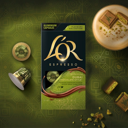 Image d'un paquet de capsules de café L'OR Espresso saveur Dubai Chocolate Style, entouré de capsules en aluminium de couleur dorée et verte, et de morceaux de chocolat aux pistaches sur un fond vert avec des motifs géométriques arabes.