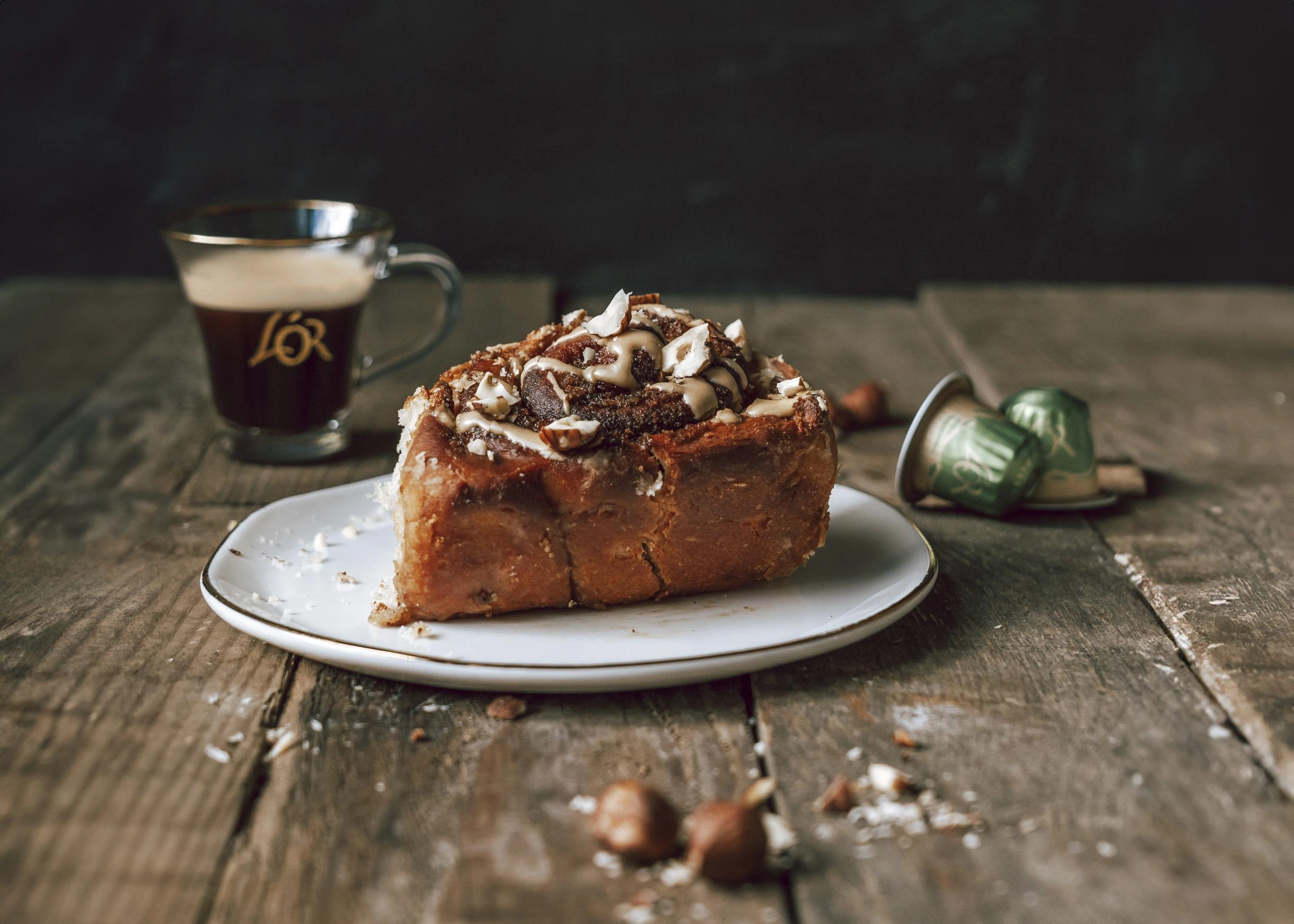 A cinnamon roll with nuts sits invitingly on a wooden table, accompanied by a steaming cup of rich L'OR Espresso or perhaps some other delightful variant from the L'OR range. The warm, aromatic coffee perfectly complements the sweetness and crunch of the pastry, creating an ideal pairing for any coffee enthusiast.