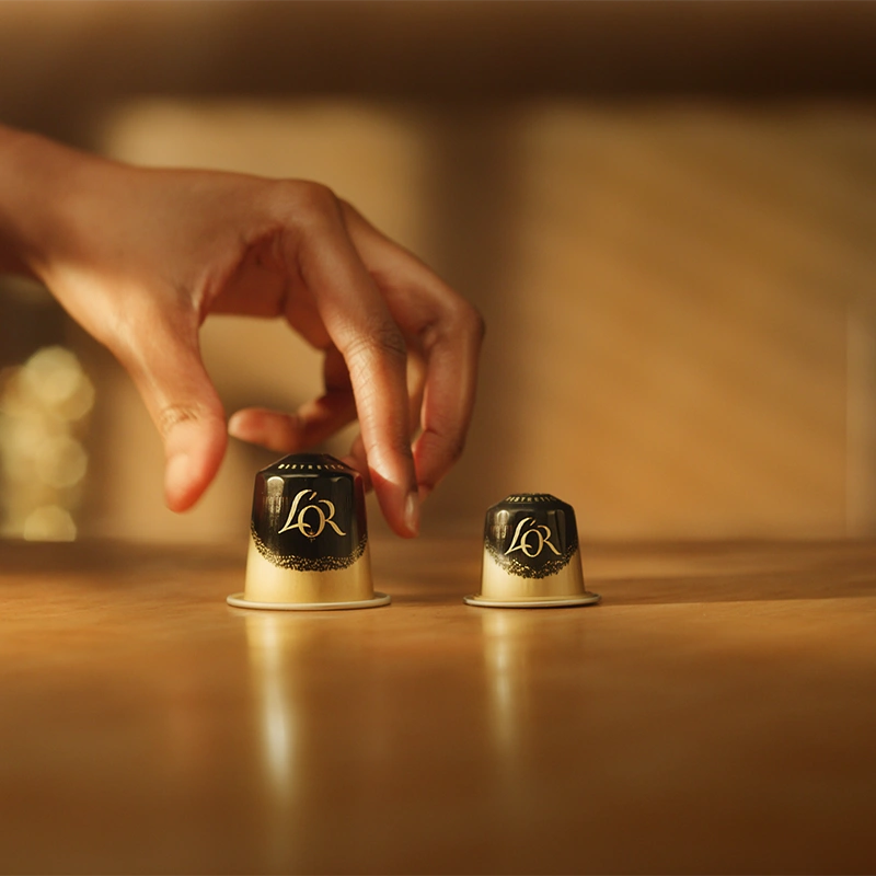 Dos cápsulas L'OR sobre una mesa dorada: una Ristretto clásica y una XXL Ristretto. Una mano toma suavemente la cápsula XXL.