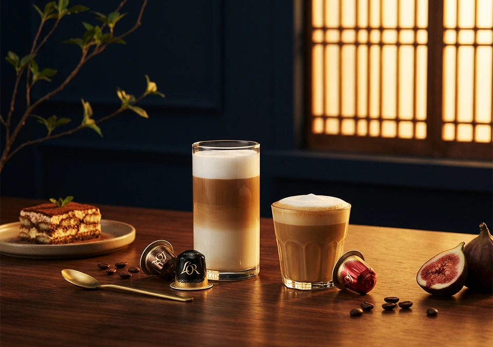 Dos bebidas de café, incluyendo latte y café con leche, servidas en vasos transparentes sobre una mesa con cápsulas