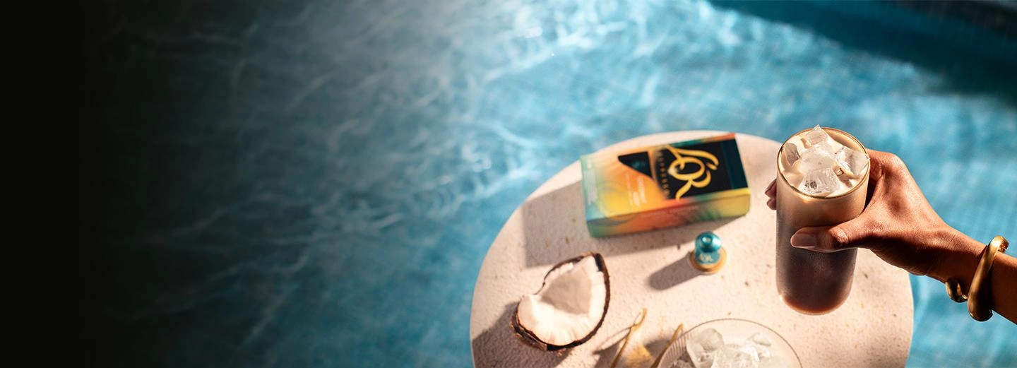 Iced coffee in a glass held above a sunlit table with L’OR coffee capsules and coconut, next to a swimming pool