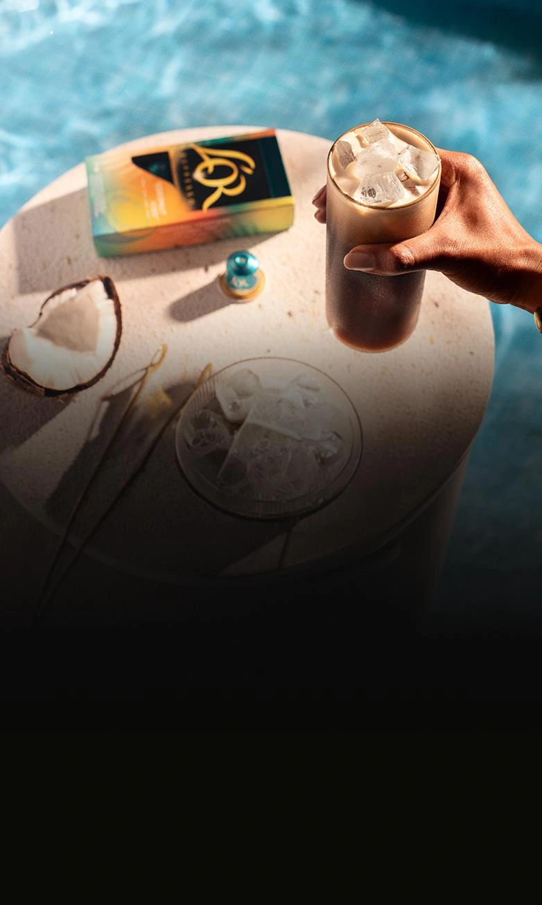 Iced coffee in a glass held above a sunlit table with L’OR coffee capsules and coconut, next to a swimming pool