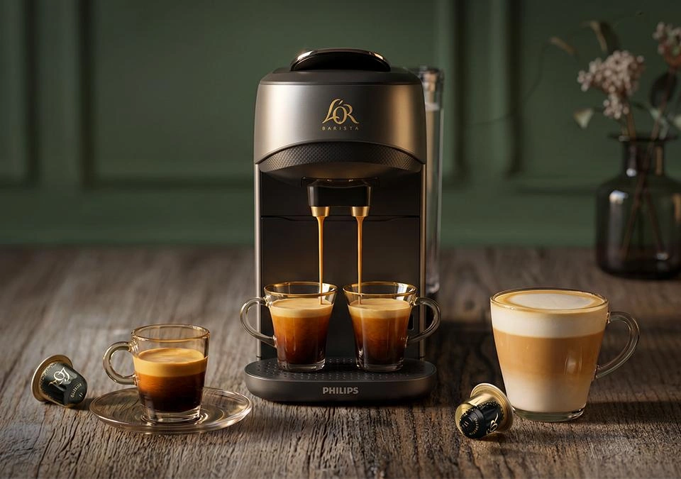 L'OR Barista coffee machine by Philips in metallic grey on a wooden table, brewing two espressos into glasses below the spout, with a cappuccino on the right, loose coffee capsules, and a vase of dried flowers against a green panelled wall.