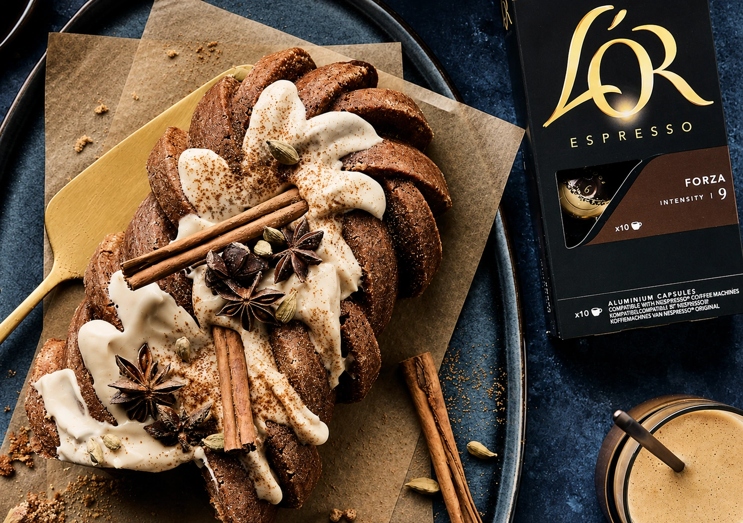 Dirty chai latte cake met spiraalvorm, afgewerkt met romige glazuur, kaneelstokjes, steranijs en kardemom, op bakpapier naast een doosje L'OR Espresso Forza capsules en een glas koffie met melk.