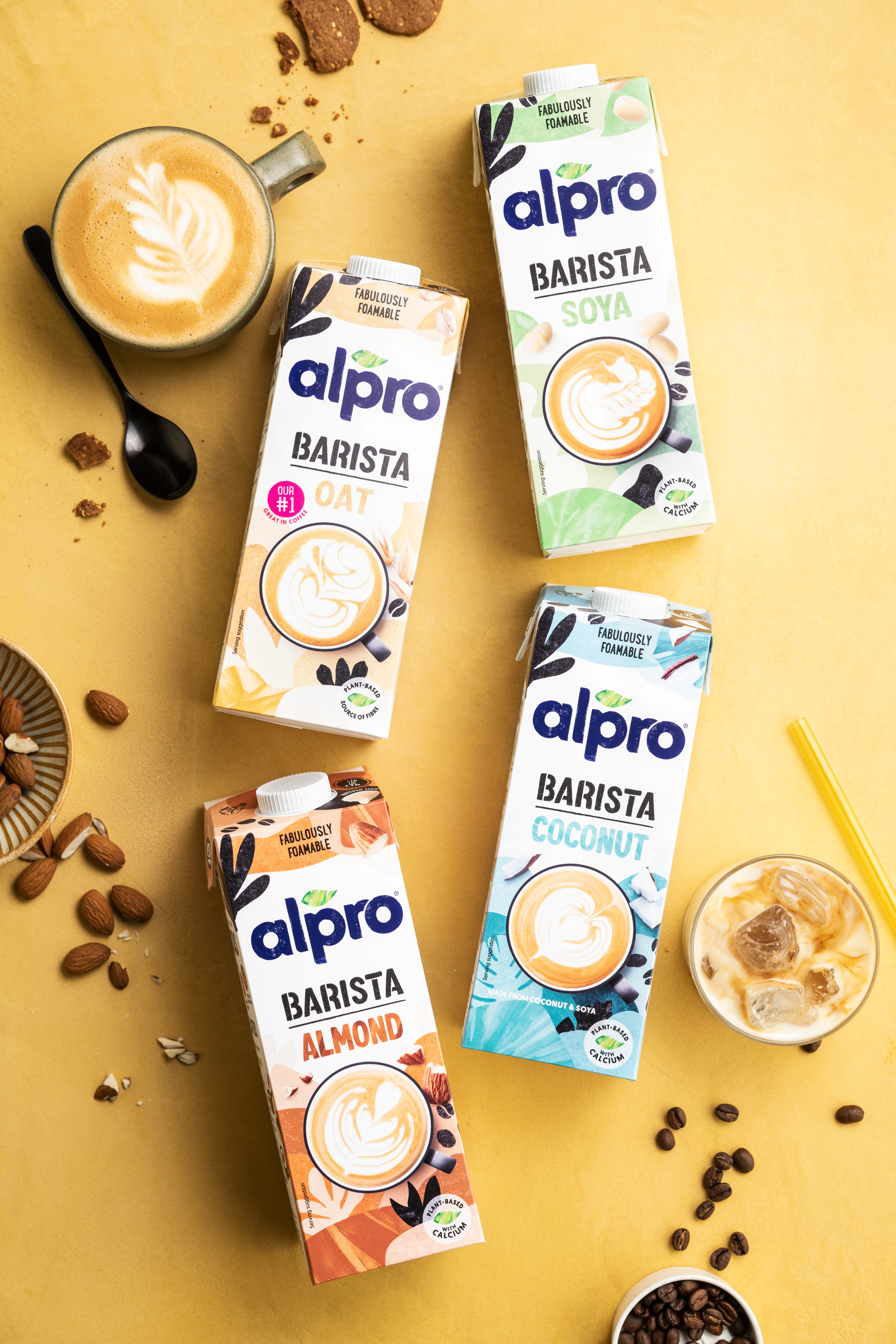 Alpro Barista oat, soya, almond and coconut drinks displayed with hot and iced coffee on a yellow background.