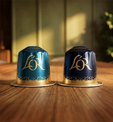 Two L'OR decaf coffee capsules, one in teal and one in dark blue with gold lettering, on a wooden table.