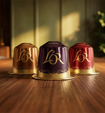 Three L'OR espresso coffee capsules in copper, purple and red, arranged together on a wooden table.