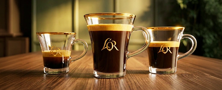 Three glass cups of L'OR coffee in different sizes, each marked with the L'OR logo, on a wooden table.