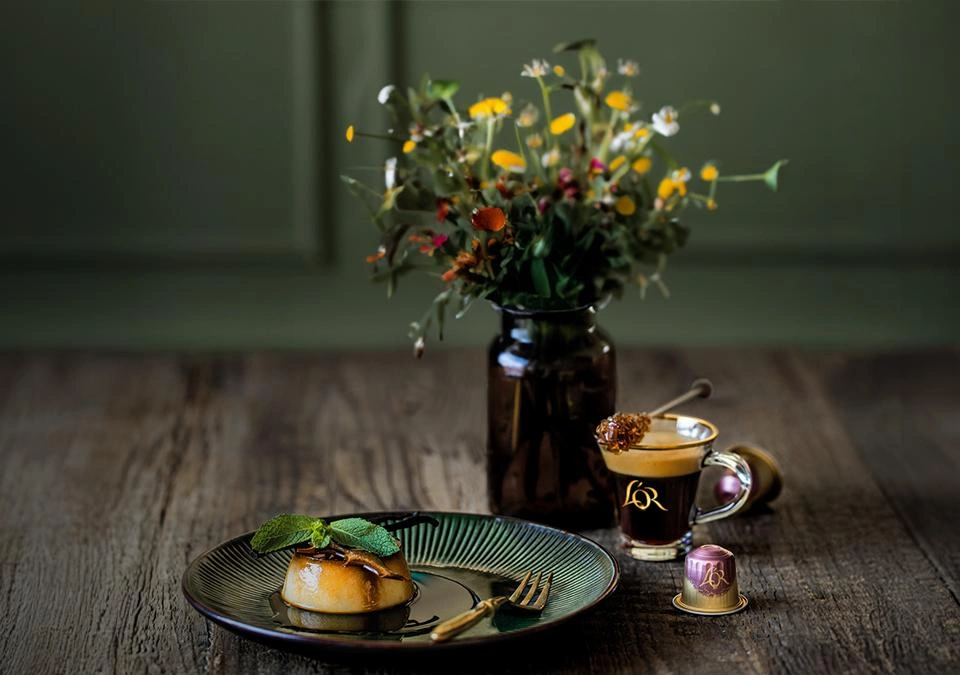 A caramel flan plate next to a L'OR Espresso Coffee cup, a coffee capsule, and a flower vase