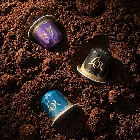 Three L'OR coffee capsules resting on freshly ground coffee.