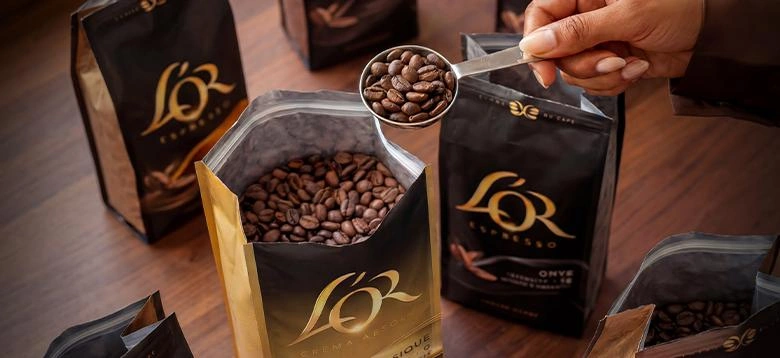 Open L’OR coffee bags with a hand holding a scoop of roasted beans.