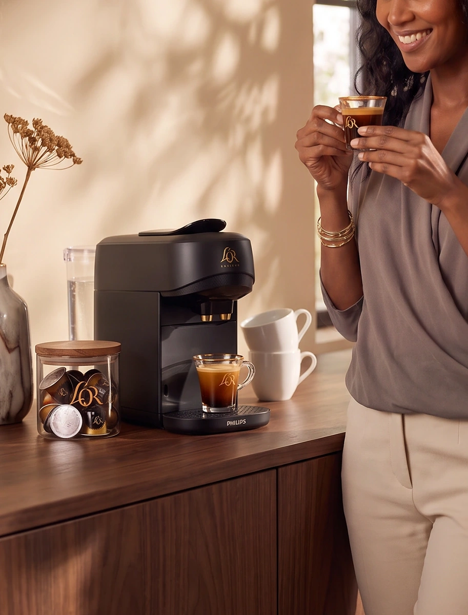 Woman standing beside a black L'OR coffee machine on a wooden sideboard.