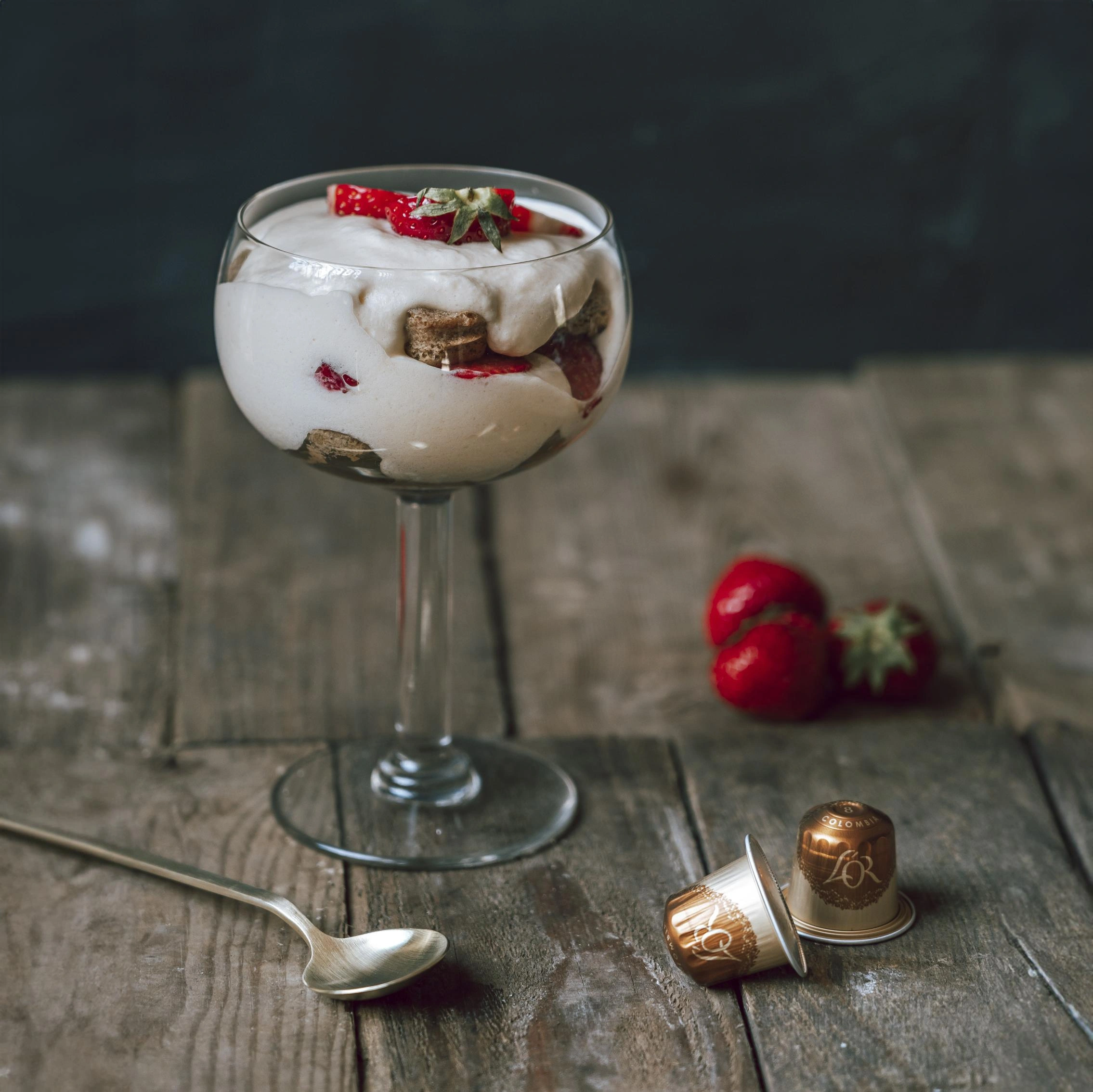Tiramisu in a glass, elegantly layered with fresh strawberries and enriched by L'OR Espresso, sits invitingly on a rustic wooden surface. Adorned with coffee capsules, this dessert offers a harmonious blend of flavors and textures.
