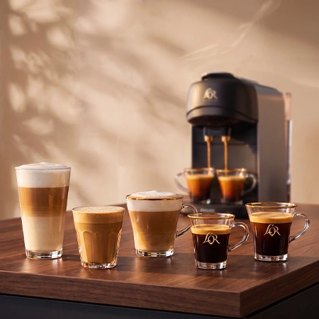Black L'OR coffee machine pouring coffee, with a latte, espresso glasses and capsules on a wooden table.